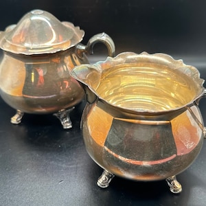 May include: A silver sugar bowl and creamer set with ornate detailing and three feet on each piece.
