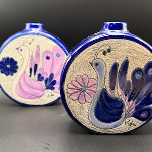 May include: Two ceramic vases with a blue rim and a beige center. Each vase features a hand-painted peacock design in shades of blue, purple, and pink. The peacock is surrounded by a floral design. The vases are likely decorative items.