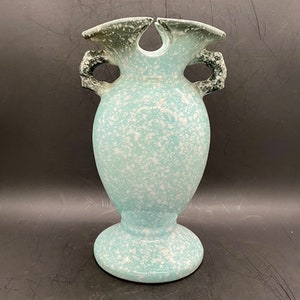 May include: A light blue ceramic vase with white speckles and two black handles. The vase has a wide mouth and a round base.