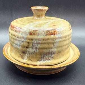 May include: A ceramic butter dish with a lid, featuring a speckled tan and brown glaze. The dish sits on a matching plate, creating a cohesive and rustic aesthetic. The lid has a small handle on top.