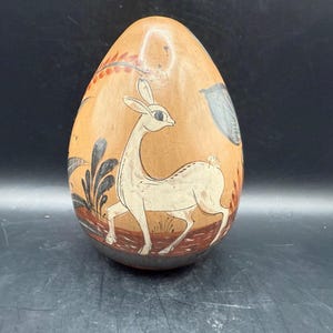 May include: A hand-painted, egg-shaped decorative item. The tan-colored surface features a white deer illustration with black and red accents. The deer is set against a backdrop of stylized plants and a blue shape.