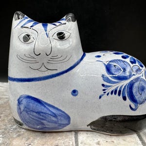 May include: A ceramic cat figurine in a seated position. The cat is primarily white with blue accents, including stripes on the ears, a collar, and floral patterns on the side. The eyes are black, and the tail is black. The figurine is approximately 8 inches long.