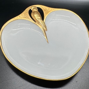 May include: A white porcelain dish with a gold rim and a gold bird design. The dish is shaped like a leaf and has a raised edge.