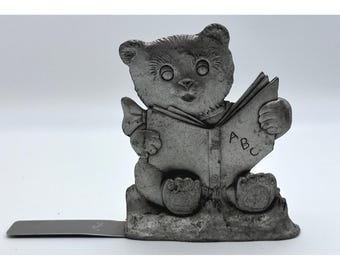 Könnte beinhalten: Zinnfarbene Bärenfigur, die ein Buch liest, auf dessen Seite "ABC" zu sehen ist. Der Bär sitzt und hat eine kleine Fliege. Ein flaches, rechteckiges Metallstück erstreckt sich von der Basis, wahrscheinlich als Buchstütze.