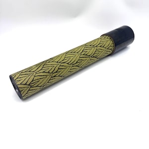 May include: A cylindrical kaleidoscope featuring a long body wrapped in a textured olive green paper with a repeating black leaf pattern. The device has a smooth, dark brown or black circular eyepiece at one end and a clear lens at the other end.