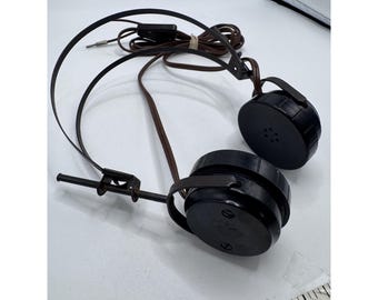 May include: Vintage black headphones with circular ear cups and a brown cord. The headphones have a metal frame and a plug for connecting to a device. The ear cups have small holes. The headphones are on a white surface.