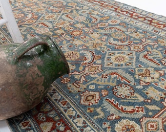 May include: A woven area rug featuring a complex geometric and floral pattern in shades of blue, rust red, and cream. Beside the rug sits a large, weathered ceramic jug with a green glazed upper section and a textured, earthy brown base.