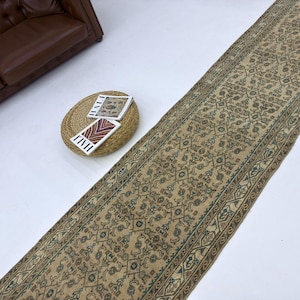 Könnte beinhalten: Ein langer, rechteckiger beigefarbener Teppich mit einem komplizierten Blumenmuster und einem braunen Rand. Der Teppich liegt auf einer weißen Oberfläche, mit einem braunen Ledersofa und einem kleinen runden gewebten Tisch mit Zeitschriften.