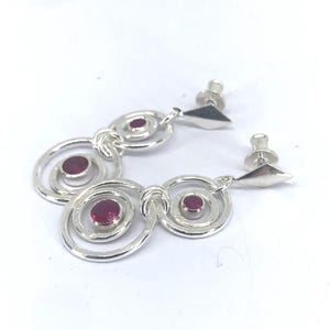 May include: A pair of silver drop earrings featuring a series of interconnected circular rings. Each earring is accented with a small, round, deep pink gemstone set in the centre of the circles. The earrings include diamond-shaped posts with backs.