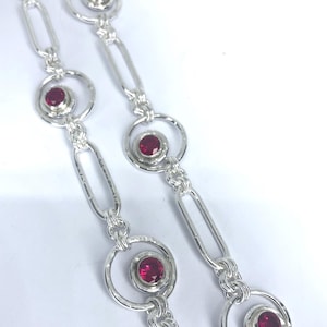 May include: A silver-toned chain necklace featuring alternating elongated oval links and circular links. Each circular link contains a small, round, faceted deep red gemstone set in the centre. The metal surface has a textured, hammered finish.