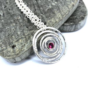 May include: A silver pendant necklace featuring a spiral design with a textured finish. The pendant has a central, round, pink gemstone. The chain is a double strand of small, linked circles. The necklace is displayed on a gray stone.