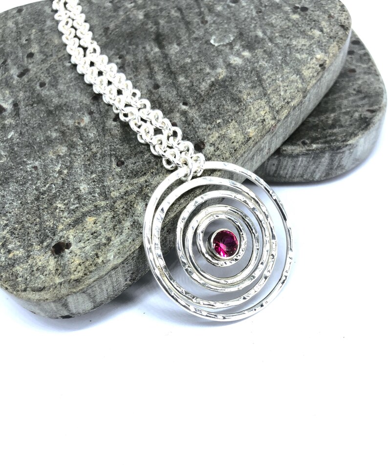 May include: A silver pendant necklace featuring a spiral design with a textured finish. The pendant has a central, round, pink gemstone. The chain is a double strand of small, linked circles. The necklace is displayed on a gray stone.