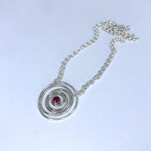 May include: A silver necklace featuring a spiral pendant with a small, round, red gemstone at the center. The pendant is connected to a delicate chain. The necklace is displayed on a white background.