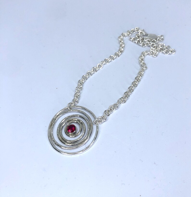 May include: A silver necklace featuring a spiral pendant with a small, round, red gemstone at the center. The pendant is connected to a delicate chain. The necklace is displayed on a white background.
