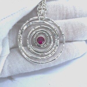 May include: A silver pendant with a series of concentric circles, each with a hammered texture. The center features a small, round, red gemstone. The pendant hangs from a silver chain.