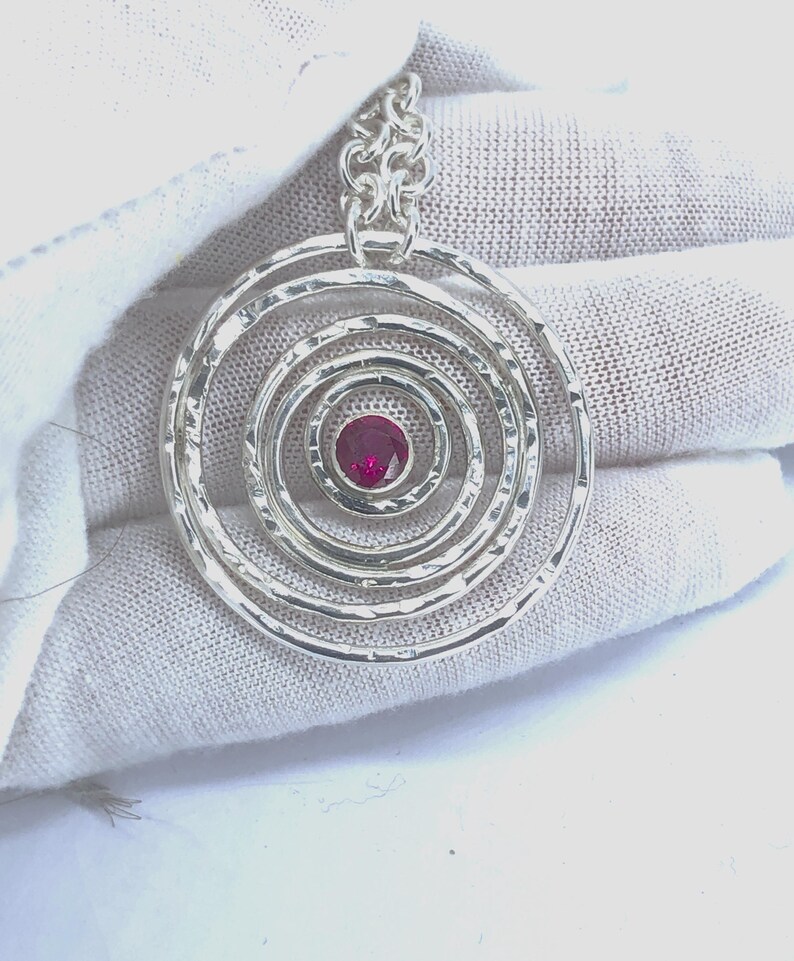 May include: A silver pendant with a series of concentric circles, each with a hammered texture. The center features a small, round, red gemstone. The pendant hangs from a silver chain.