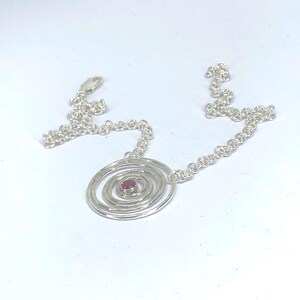 May include: A silver necklace featuring a spiral pendant with a small, round, red gemstone at its center. The pendant is connected to a delicate chain. The necklace is displayed on a white background.