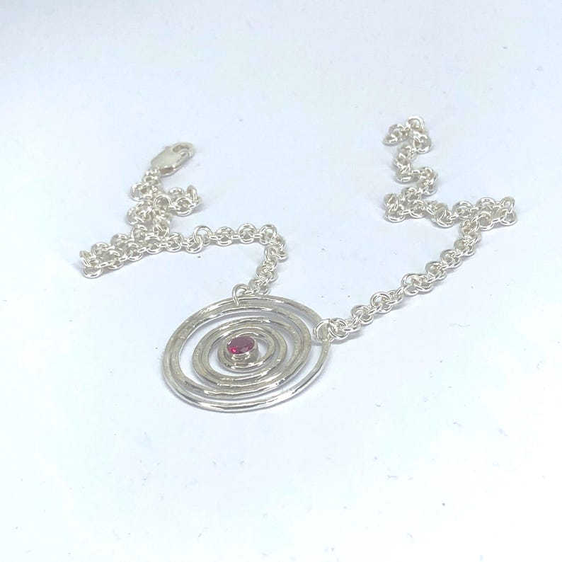 May include: A silver necklace featuring a spiral pendant with a small, round, red gemstone at its center. The pendant is connected to a delicate chain. The necklace is displayed on a white background.