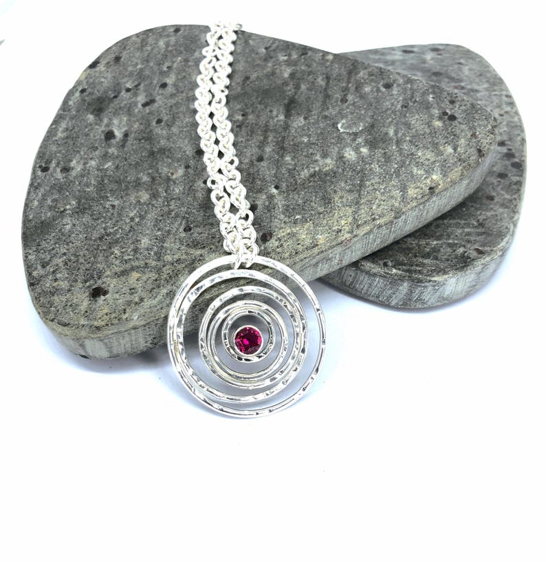May include: A silver pendant necklace featuring a spiral design with a small, round, red gemstone at the center. The pendant hangs from a delicate chain. The necklace is displayed on a gray stone surface.