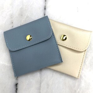 May include: Two small, square pouches. One is a muted blue, the other is cream-colored. Each has a flap closure secured by a gold-colored snap. The pouches are made of a smooth material and are likely for small items.