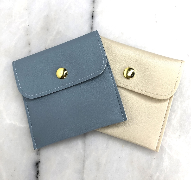 May include: Two small, square pouches. One is a muted blue, the other is cream-colored. Each has a flap closure secured by a gold-colored snap. The pouches are made of a smooth material and are likely for small items.