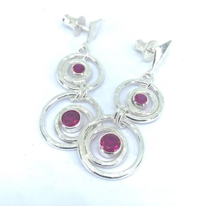 May include: A pair of silver-toned drop earrings featuring two interlocking hammered circular rings. Each ring contains a round, deep pink gemstone set in the centre. The earrings include standard post-style backs for secure attachment to the ear.