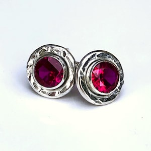 May include: A pair of silver stud earrings featuring round, deep magenta gemstones. The gemstones are set in a textured, circular frame. The earrings are shown against a white background.