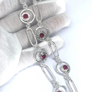 May include: A silver chain necklace featuring alternating elongated oval links and circular links. Each circular link contains a single, round, deep pink gemstone set in the centre. The metal has a bright, polished finish throughout the design.