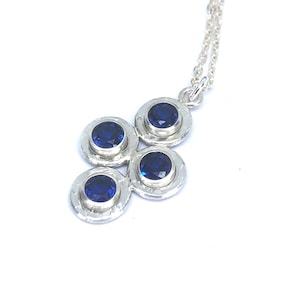 May include: A silver necklace with a pendant composed of four circular settings, each containing a deep blue gemstone. The pendant is attached to a fine silver chain. The gemstones are a vivid blue, set in a textured silver setting.