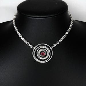 May include: A silver necklace featuring a spiral pendant with a central red gemstone. The pendant is composed of concentric circles with a textured finish. The necklace is displayed on a black mannequin.