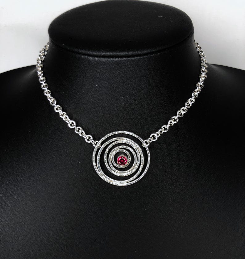 May include: A silver necklace featuring a spiral pendant with a central red gemstone. The pendant is composed of concentric circles with a textured finish. The necklace is displayed on a black mannequin.