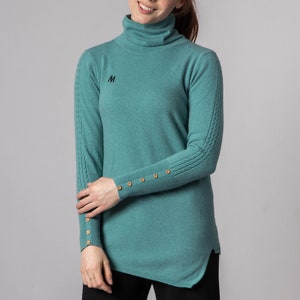 Puede incluir: Un suéter de cuello alto azul turquesa con botones dorados en las mangas. El suéter tiene un diseño de punto de cable en las mangas y una pequeña 'M' bordada en la parte delantera.