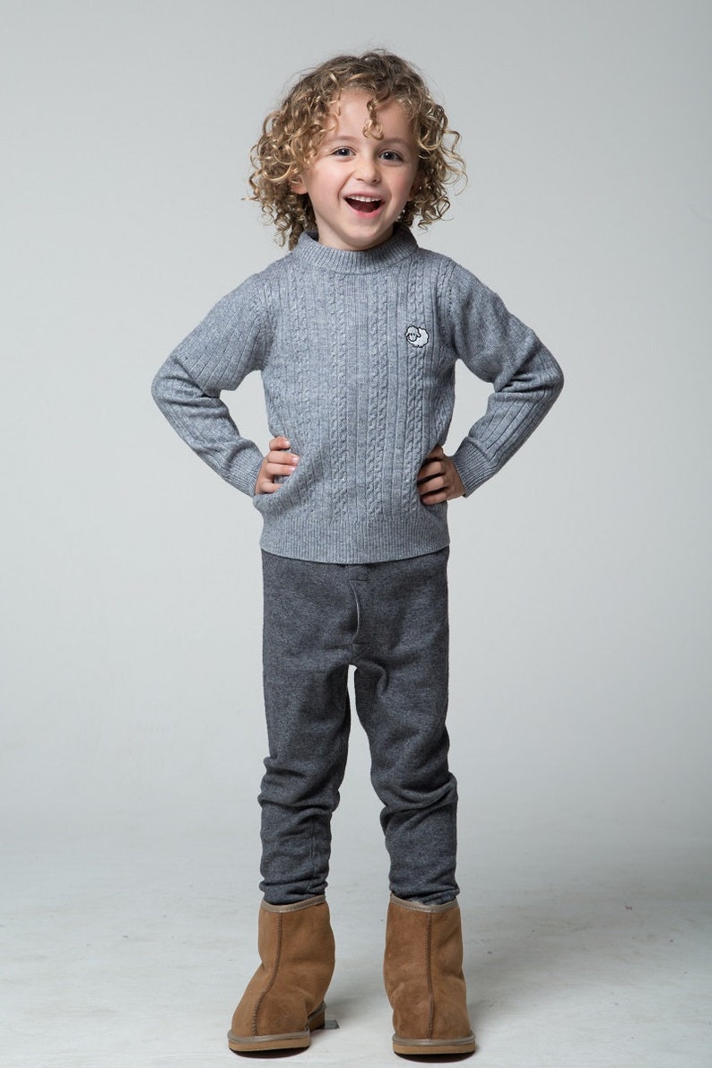 Puede incluir: Un ni&ntilde;o peque&ntilde;o con un su&eacute;ter gris de punto de cable con una peque&ntilde;a oveja bordada en la parte delantera, pantalones grises y botas de gamuza marr&oacute;n. El ni&ntilde;o sonr&iacute;e y tiene el pelo rizado.