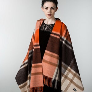 May include: A woman wearing a black dress with a lace neckline is wearing a large, orange, brown, and cream plaid shawl with fringe. The shawl has a small, white label with the word "ORVIS" on it.