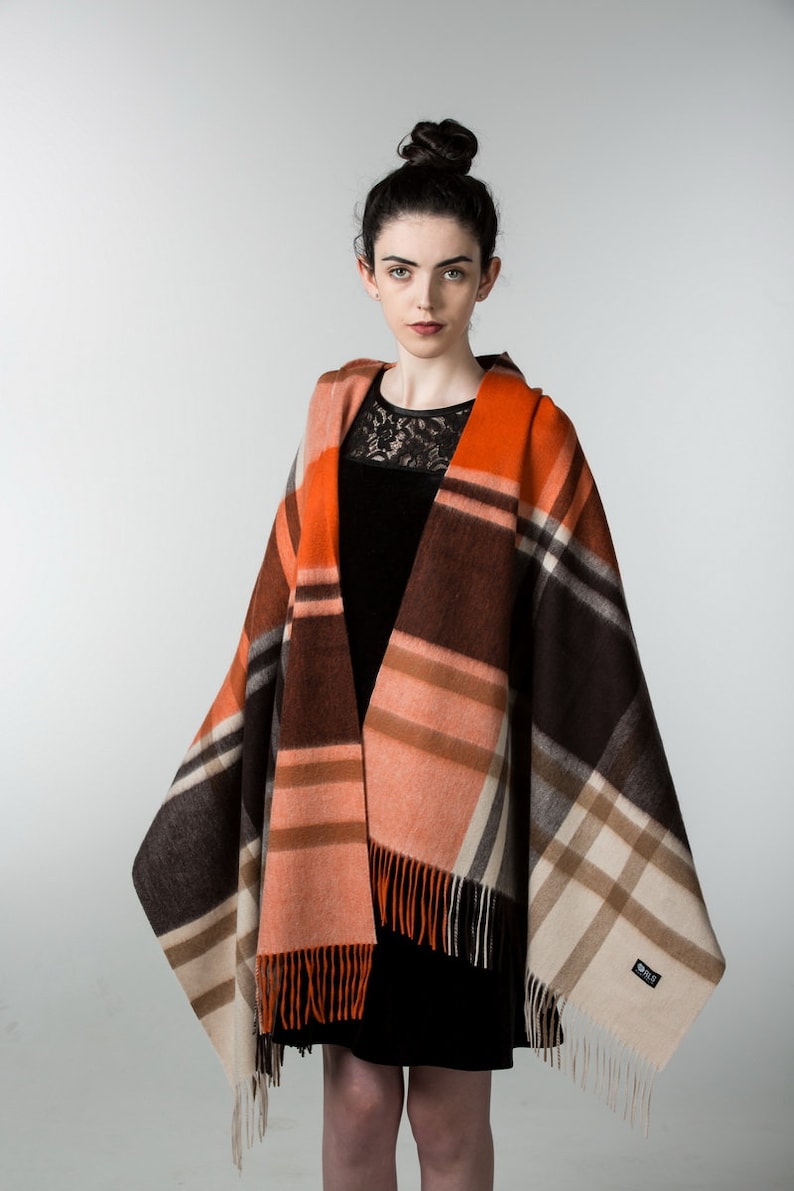 May include: A woman wearing a black dress with a lace neckline is wearing a large, orange, brown, and cream plaid shawl with fringe. The shawl has a small, white label with the word "ORVIS" on it.