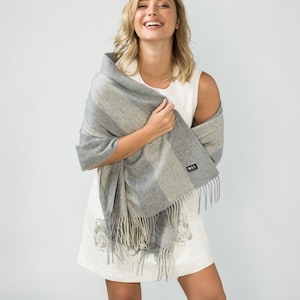 May include: A woman wearing a white sleeveless dress with a gray and white striped shawl with fringe. The shawl is draped over her shoulders and she is holding it in her hands.