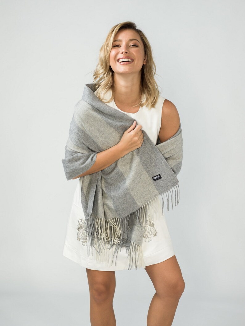 May include: A woman wearing a white sleeveless dress with a gray and white striped shawl with fringe. The shawl is draped over her shoulders and she is holding it in her hands.