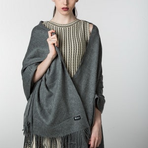 May include: A gray pashmina shawl with fringe, worn over a striped sleeveless top. The shawl has a small black label with the text "B.L.S. Collection" on it.