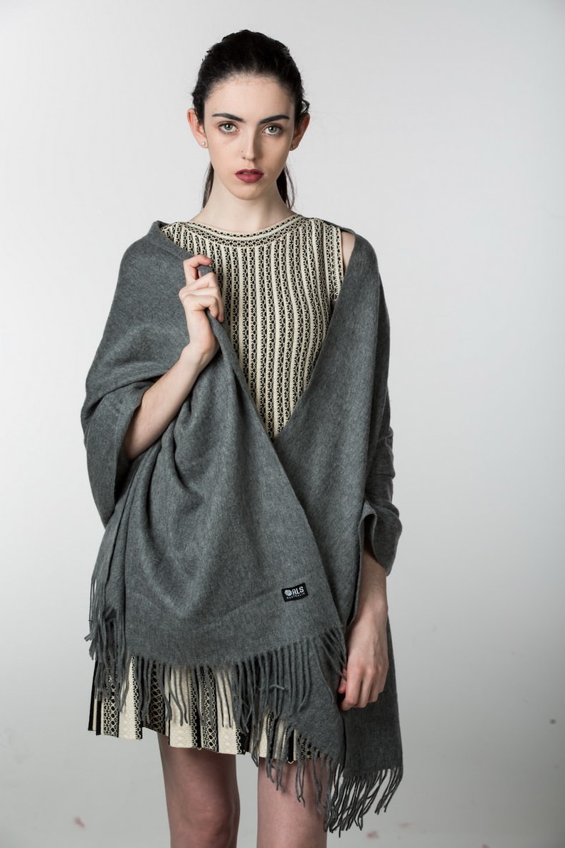 May include: A gray pashmina shawl with fringe, worn over a striped sleeveless top. The shawl has a small black label with the text "B.L.S. Collection" on it.