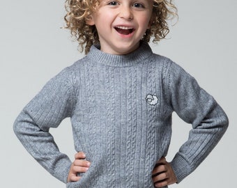 Puede incluir: Un niño pequeño con un suéter gris de punto de cable con una pequeña oveja blanca bordada en la parte delantera. El suéter tiene cuello alto y mangas largas.