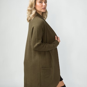 K&ouml;nnte beinhalten: Ein langer, olivgr&uuml;ner Cardigan mit Schalkragen und einer einzigen Tasche auf der linken Seite. Der Cardigan ist aus einem weichen, gestrickten Stoff gefertigt.