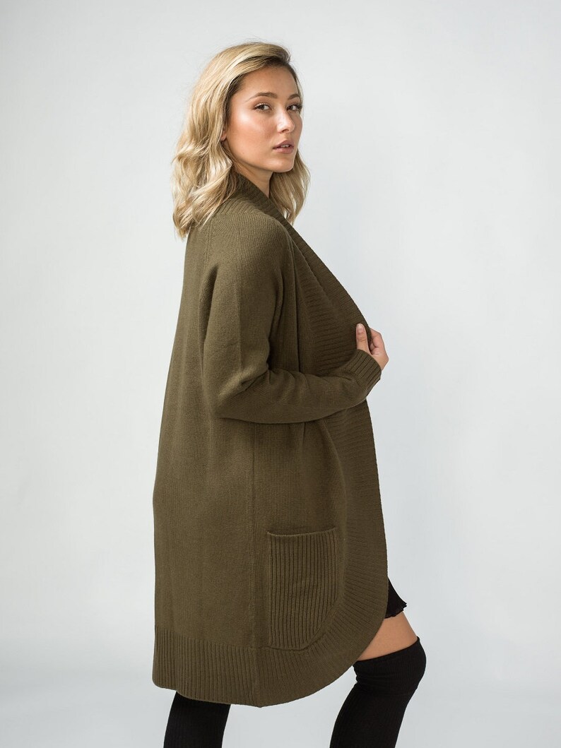 K&ouml;nnte beinhalten: Ein langer, olivgr&uuml;ner Cardigan mit Schalkragen und einer einzigen Tasche auf der linken Seite. Der Cardigan ist aus einem weichen, gestrickten Stoff gefertigt.