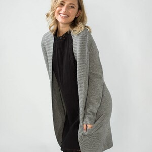 K&ouml;nnte beinhalten: Ein grauer, lang&auml;rmliger Cardigan mit Rippenstruktur und Taschen. Der Cardigan wird &uuml;ber einem schwarzen Kleid getragen.