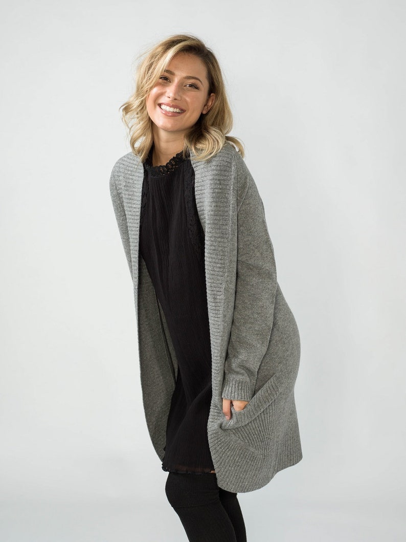 K&ouml;nnte beinhalten: Ein grauer, lang&auml;rmliger Cardigan mit Rippenstruktur und Taschen. Der Cardigan wird &uuml;ber einem schwarzen Kleid getragen.