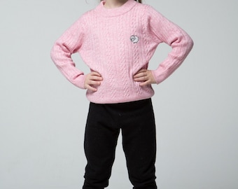 Puede incluir: Una niña pequeña con un suéter rosa con un pequeño diseño de oveja blanca, leggings negros y botas marrones. Está sonriendo y de pie con las manos en las caderas.