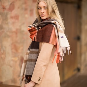 May include: A woman wearing a beige coat and a plaid scarf with brown, orange, and white stripes. The scarf has fringe on the ends and a small tag with the word "Hills" on it. She is holding a brown fedora hat.