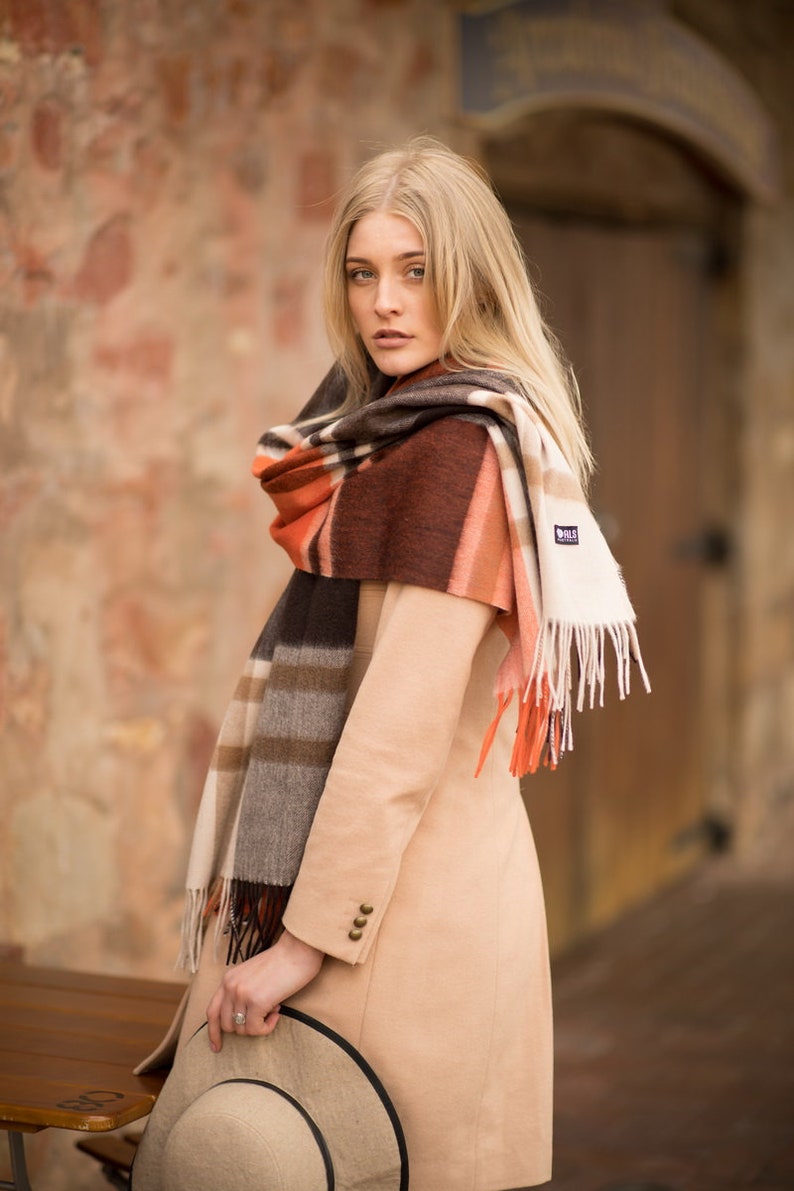 May include: A woman wearing a beige coat and a plaid scarf with brown, orange, and white stripes. The scarf has fringe on the ends and a small tag with the word "Hills" on it. She is holding a brown fedora hat.