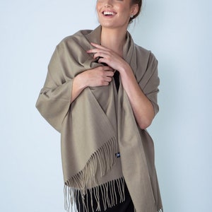 May include: A light brown shawl with fringe edges. The shawl is draped around a person's shoulders and is being held in place by their hands.