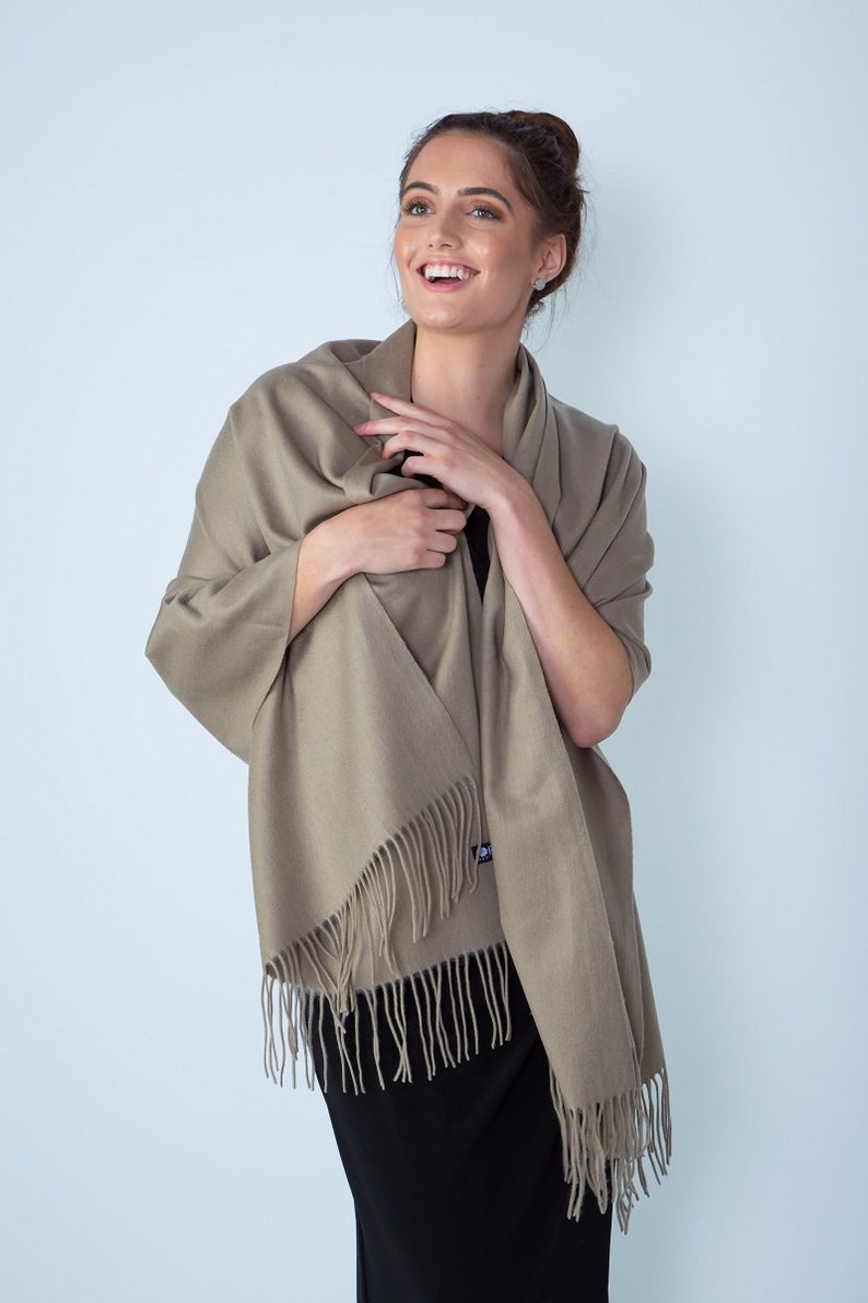 May include: A light brown shawl with fringe edges. The shawl is draped around a person's shoulders and is being held in place by their hands.