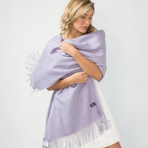 May include: A lavender pashmina shawl with fringe. The shawl is draped over a person's shoulders and is held in place by their hands.